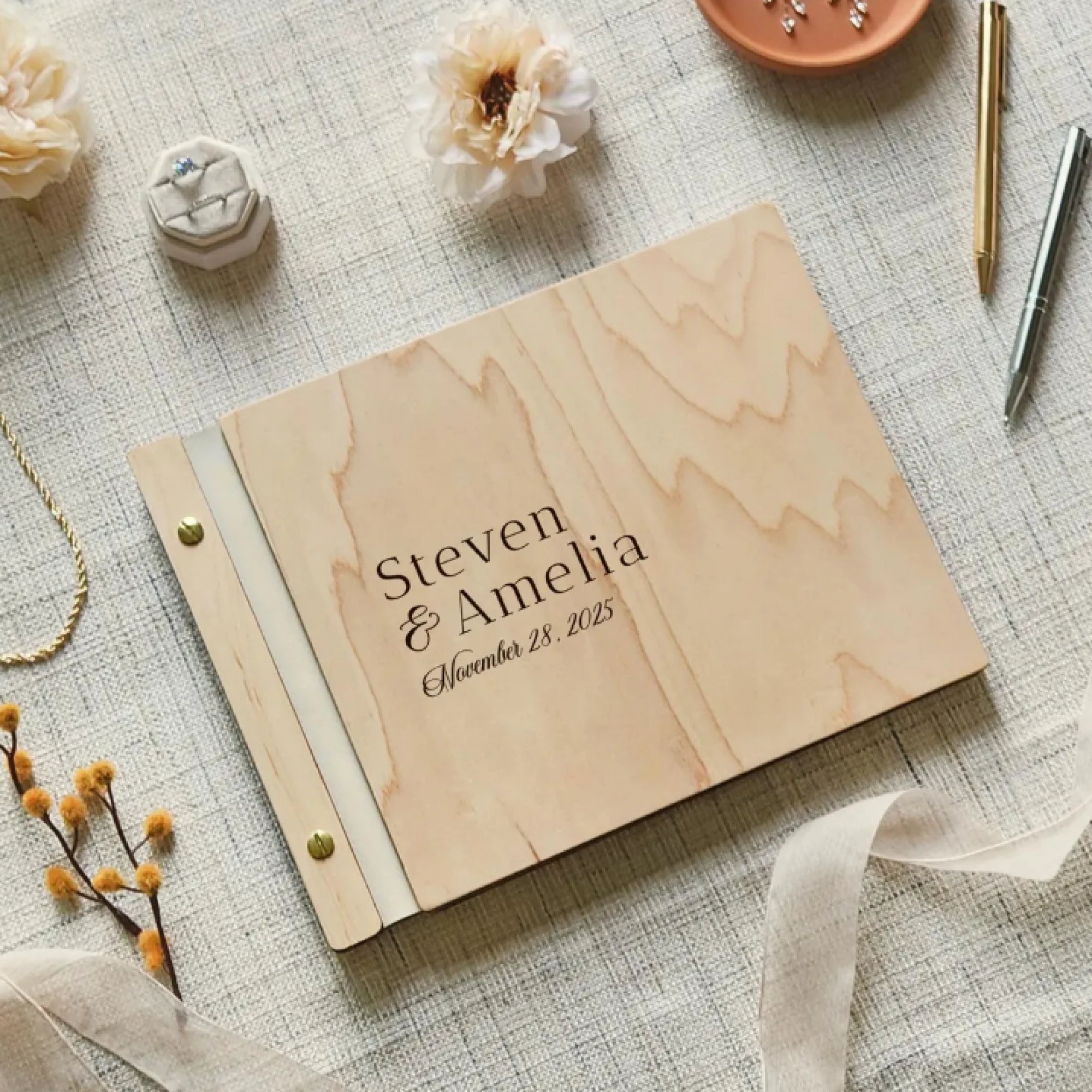 Wooden photo album guestbook cover engraved with the couple's names, laid out with flowers and a pen