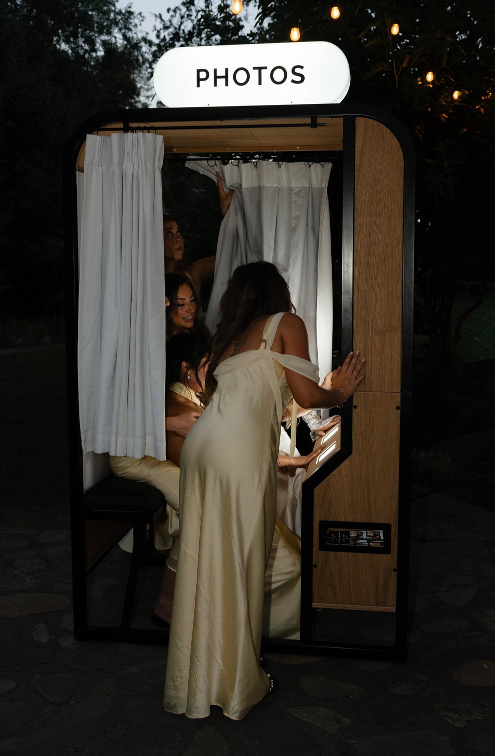 A guest in a gold dress stepping into the enclosed vintage photo booth under its PHOTOS sign