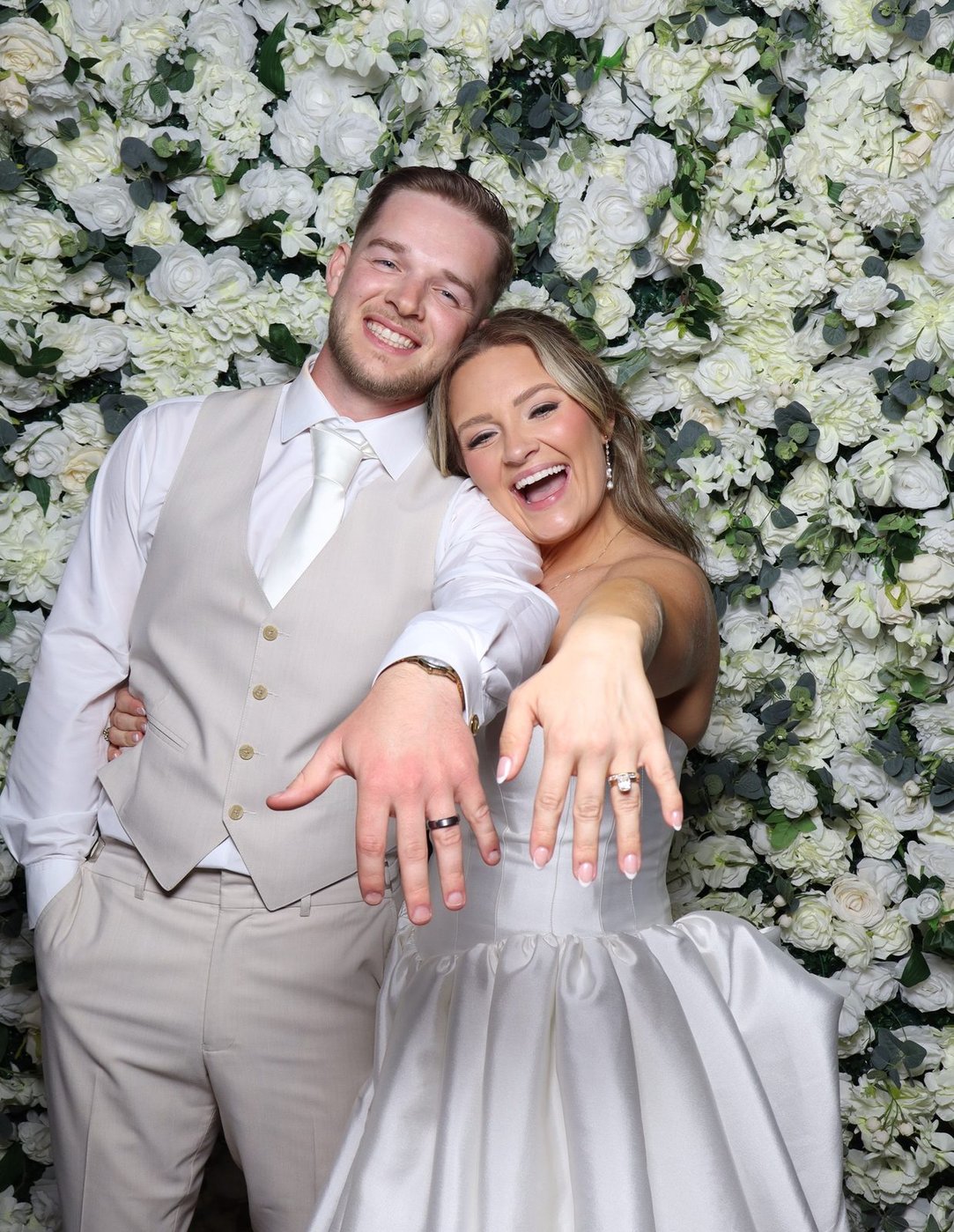 A newly married couple showing their rings at the flower wall