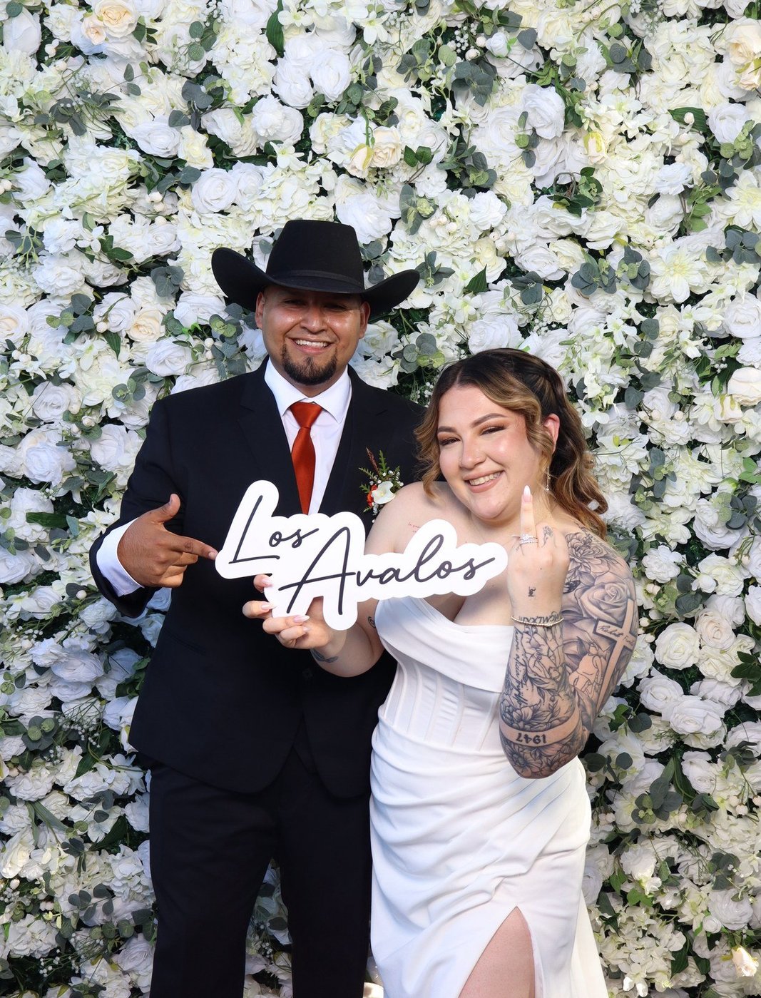 A couple holding a custom Los Avalos name sign in front of the white flower wall