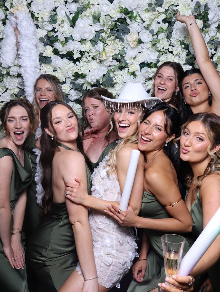 Bridesmaids in sage dresses crowding around the bride at the flower wall