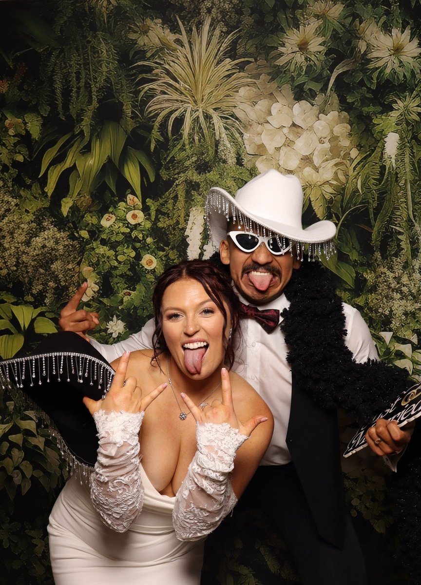 A couple in cowboy hats and a feather boa in front of a greenery backdrop