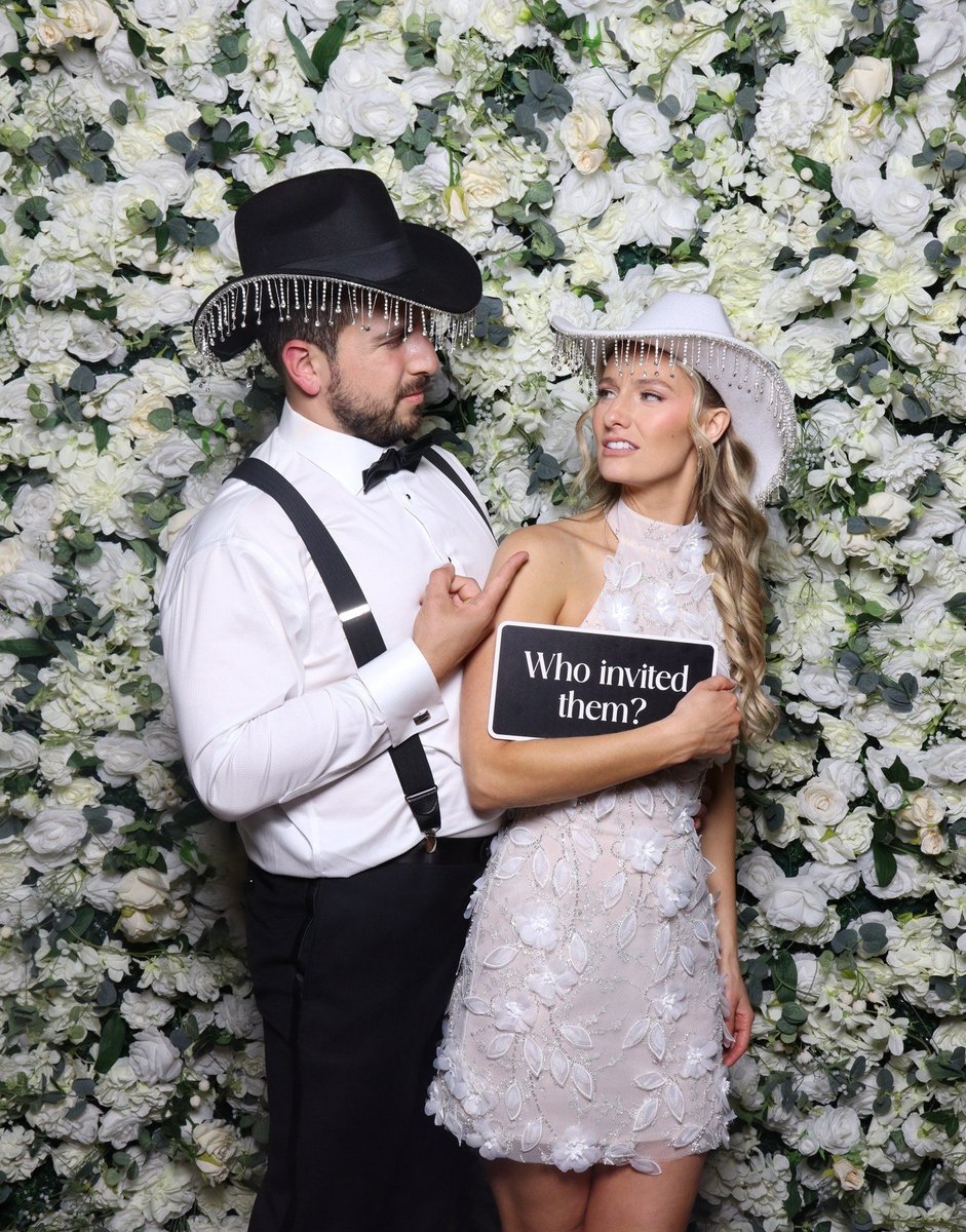 A couple in cowboy hats holding a Who Invited Them sign at the flower wall