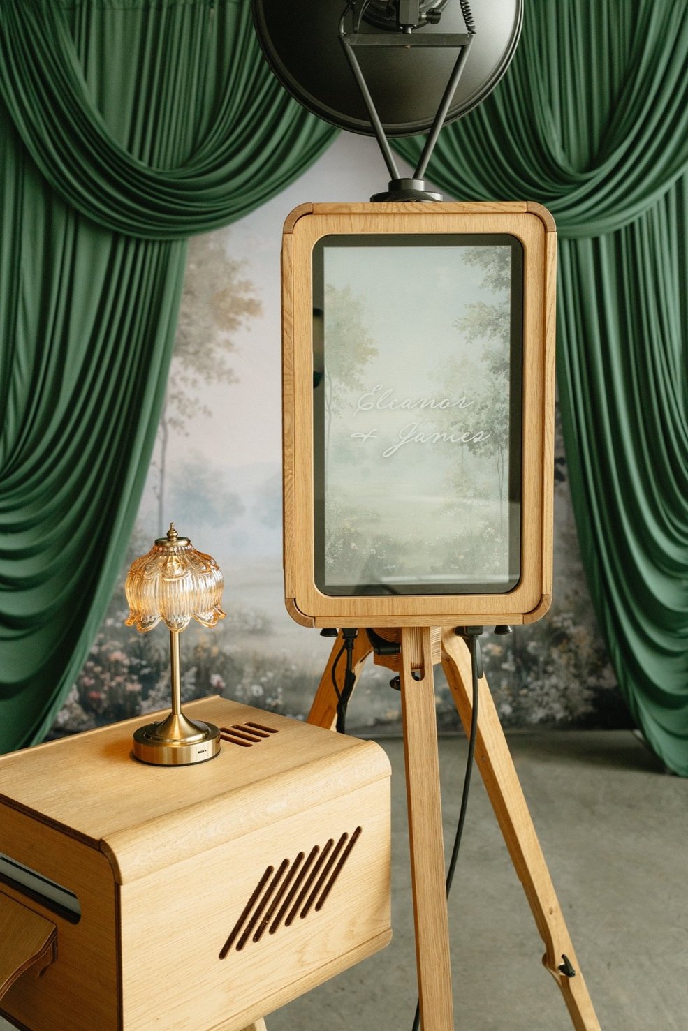 The wooden photo booth on its tripod in front of green velvet drapes and a forest backdrop